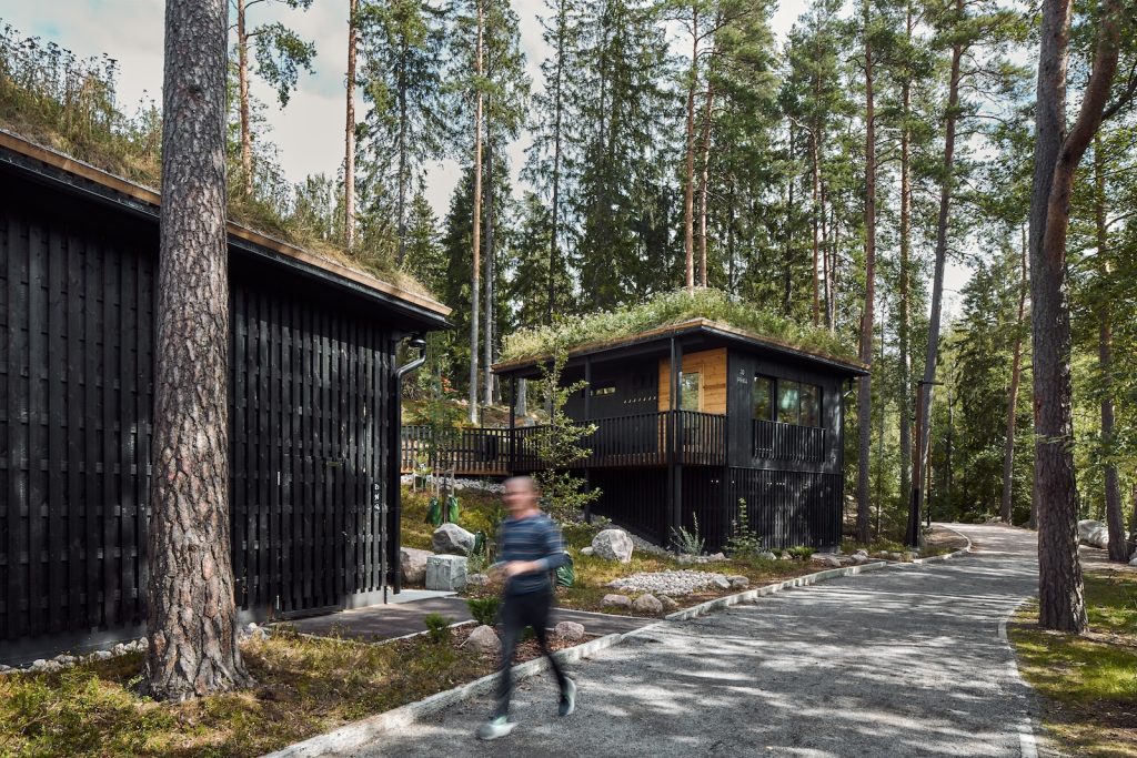 Woman jogging on a path in a forest with two cabins on the left-hand side of the image.