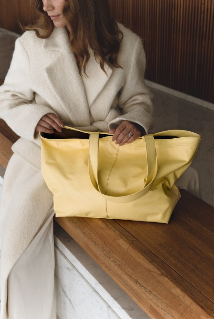 Nainen jolla on valkoinen takki istuu penkillä ja hänellä on edessään keltainen laukku