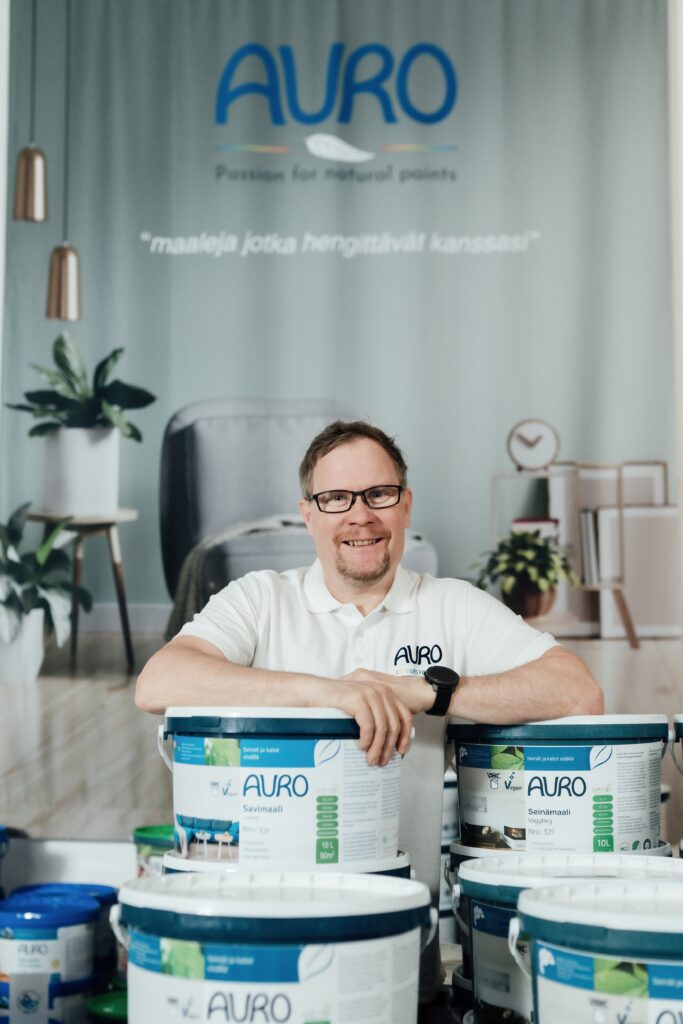 Suomen Luonnonmaalien perustaja katsoo kameraan maalipurkkien takana seisten.