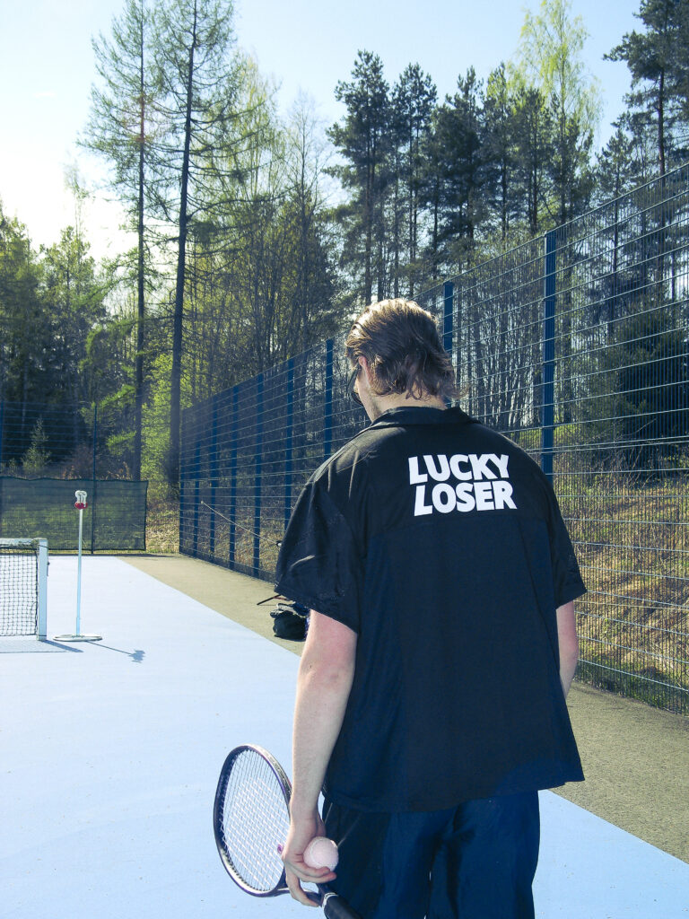Mies kävelee pois päin kamerasta ja hänellä on vasemmassa kädessä tennismaila